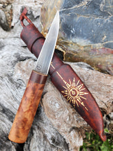 Heritage Puukko knife with wood handle and its leather sheath embossed with a gold Northern Star compass. It is displayed on a natural textured surface