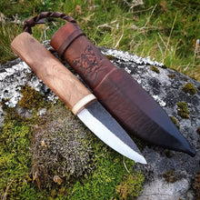 The Homestead Puukko with a wood and bone handle, displayed with a custom leather sheath, placed on a rock with green moss.