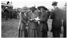 Photograph of three ladies at the Masterton Racing Club's meeting at the Opaki Race Course in 1920. A large crowd of race-goers is in the background.