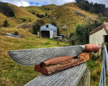Off Track Machete with kanuka handle and Damascus steel on a totara display stand, Tararua Ranges and farm sheds behind.