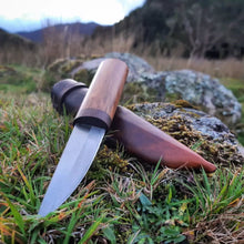 Puukko knife with a polished blade and contrasting woods in the handle, resting on its custom leather sheath in a grassy farm paddock