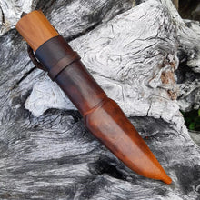 Bushcraft puukko in its leather sheath, against a backdrop of weathered driftwood.