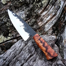 A hand forged blade reflects the light, the redwood handle has a lightning bolt running through the grain. The knife rests on a background of worn gnarly wood.