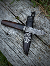 Decorative knife with a dark wood handle and damascus blade on a black leather sheath embossed with a silver rune. the knife ress on a rustic wooden surface.