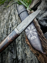 Damascus Puukko knife with a wooden handle and patterned steel blade on leather sheath, resting on a textured wooden surface.
