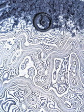 Close-up of a damascus steel surface with concentric circular patterns and our compass center disc logo stamped into the textured surface