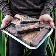 Benjamin holding a tray with stabilised redwood blocks ready to be carved into handles, standing against a flax bush