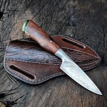 Integral damascus hunting knife with a wooden handle and pounamu pommel. It rests on its custom ambidextrous sheath, embossed with the same damascus pattern as on the blade.
